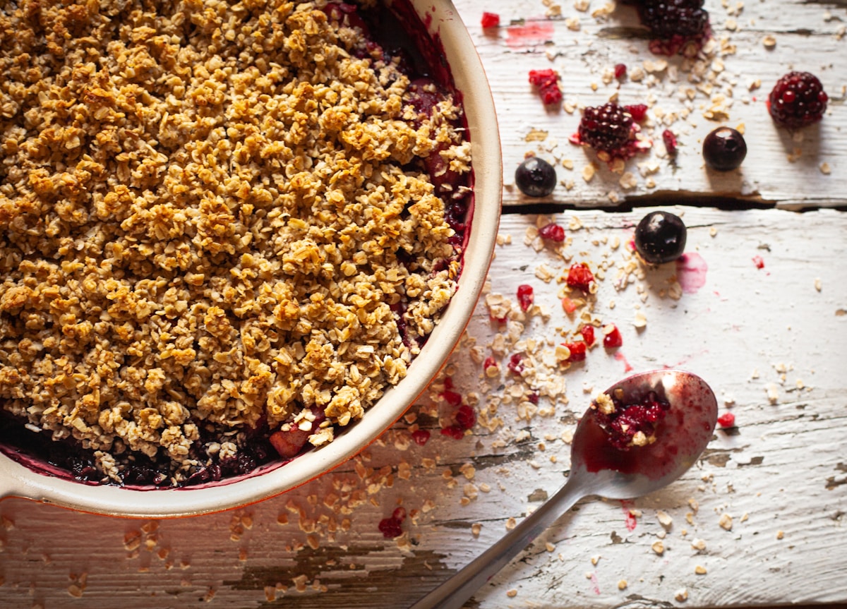 Beeren-Crumble in der Backform mit goldbraunem Haferstreu – frische Brombeeren und Beeren darunter, Holzlöffel daneben