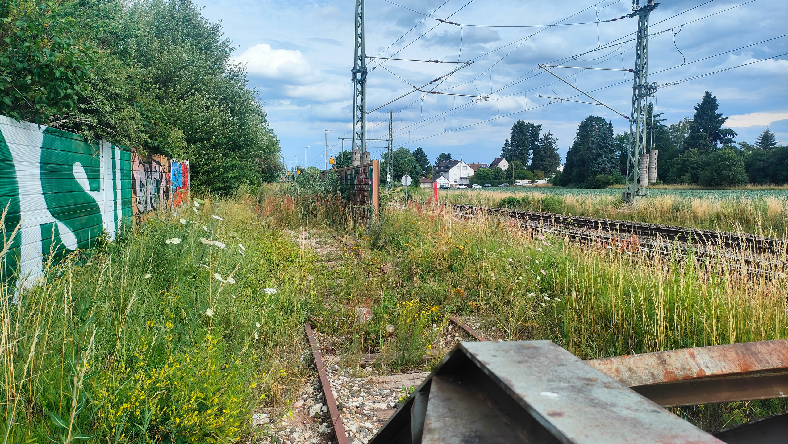 Blick zurück auf den begehbaren Weg, links eine Trennwand mit Graffiti, rechts die aktive Bahnstrecke mit Oberleitung