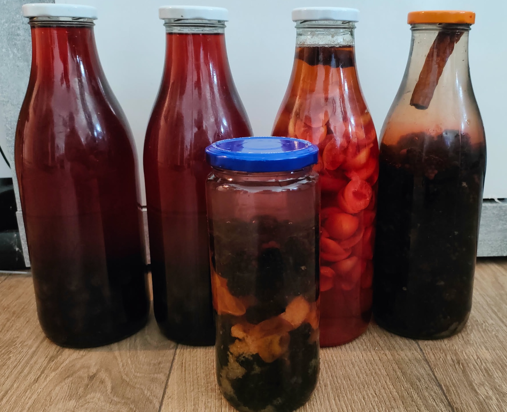Fünf Likör-Ansätze nebeneinander: zwei dunkelrote Schlehenliköre, ein kleines Glas mit blauem Deckel, ein Kirschpflaumenlikör mit sichtbaren Fruchtstücken und der neue Brombeerlikör mit Zimtstange