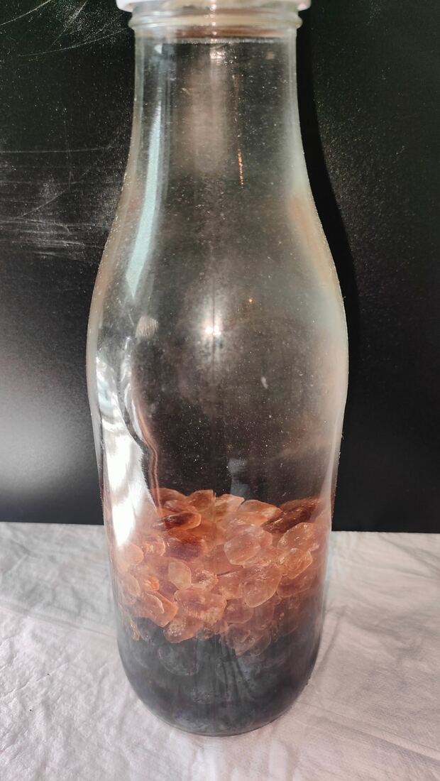 Angesetzter Schlehenlikör in einer randvollen Flasche: Schlehen am Boden, darüber brauner Kandis, bis zum Rand mit klarem Doppelkorn aufgefüllt
