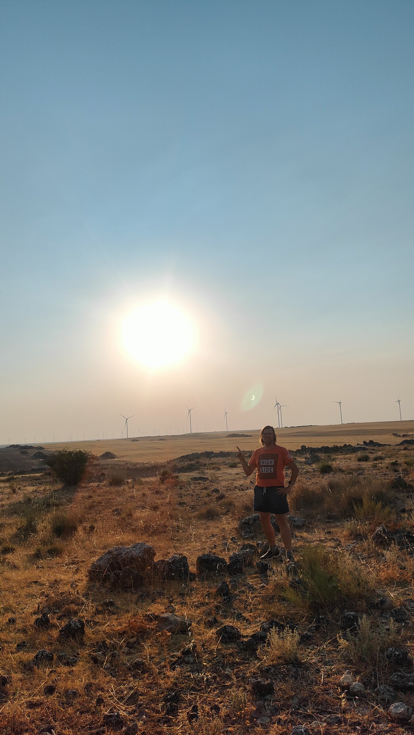 Christian steht in einem trockenen Feld mit Windrädern im Hintergrund, die große Sonne tief am Himmel, und zeigt mit der Hand eine Zwei