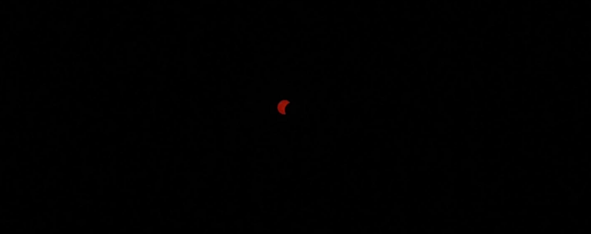Partielle Phase einer Sonnenfinsternis, durch ein Teleobjektiv fotografiert: eine schmale rote Sichel vor komplett schwarzem Himmel