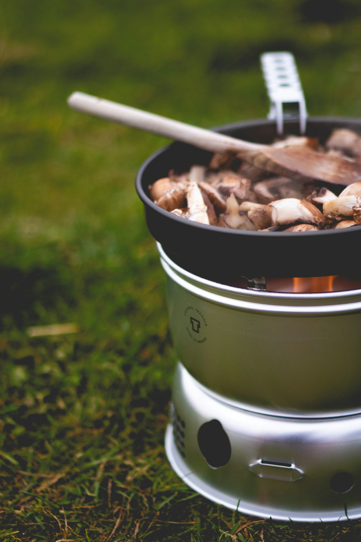 Camping-Gaskocher auf Gras mit Pilzen in der Pfanne und sichtbarer Flamme
