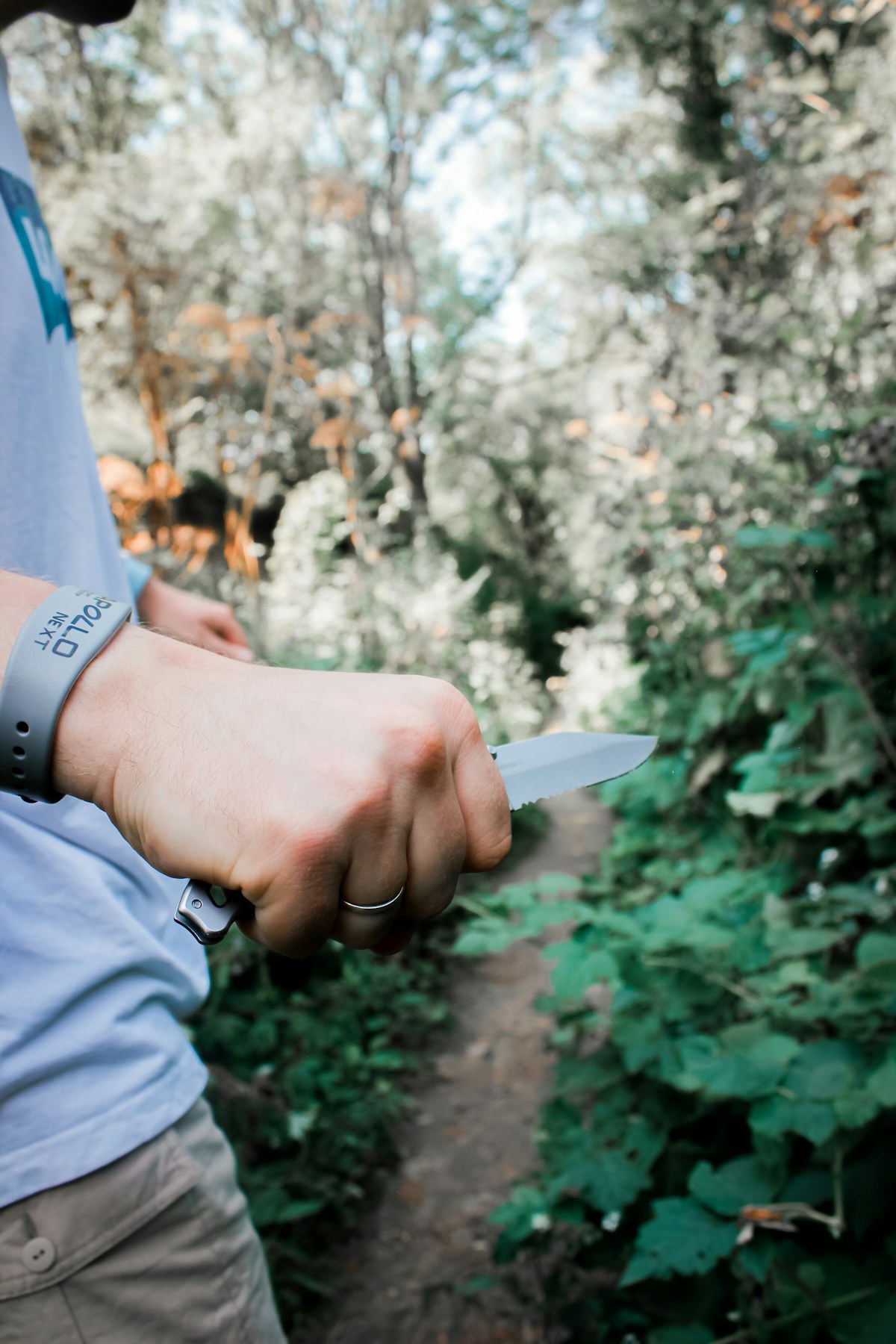 Person hält ein Outdoor-Messer mit kurzem Griff auf einem Waldweg zwischen grüner Vegetation