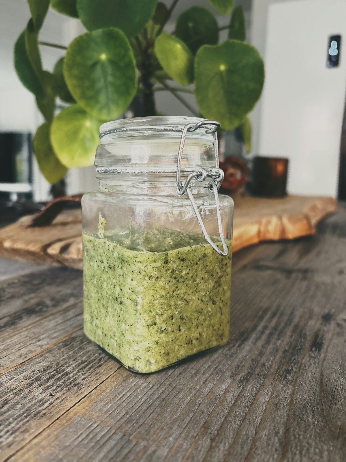 Grünes Pesto im rustikalen Einmachglas auf Holzbrett, Zimmerpflanze im Hintergrund