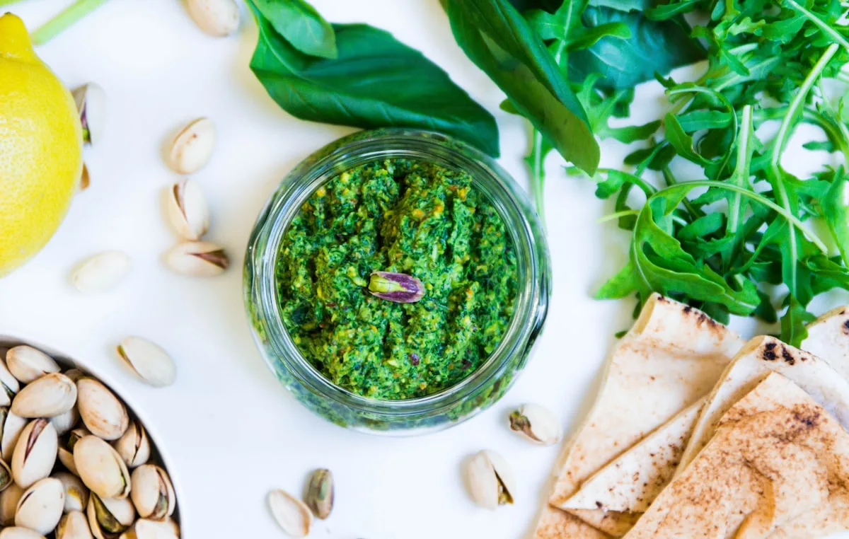 Grünes Pesto im Glasgefäß, umgeben von Nüssen, Rucola und Brot