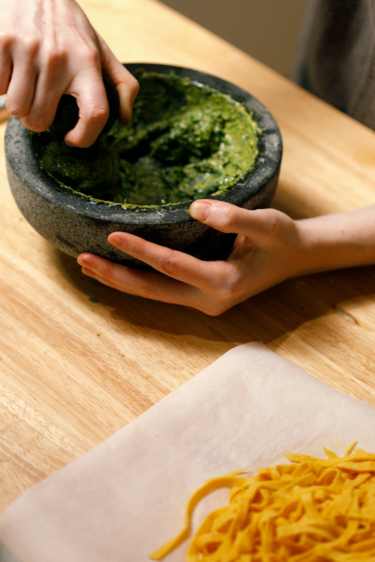 Hände reiben frisches grünes Pesto im Steinmörser, daneben frische Pasta auf Holzbrett